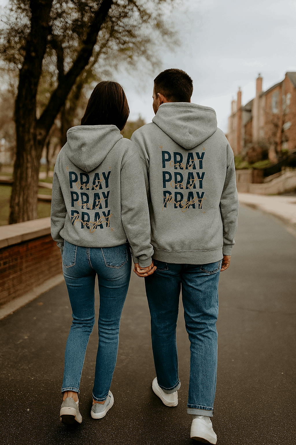 Couple wearing gray Pray On It Faith Hoodies with repeated "PRAY" text walking and holding hands outdoors
