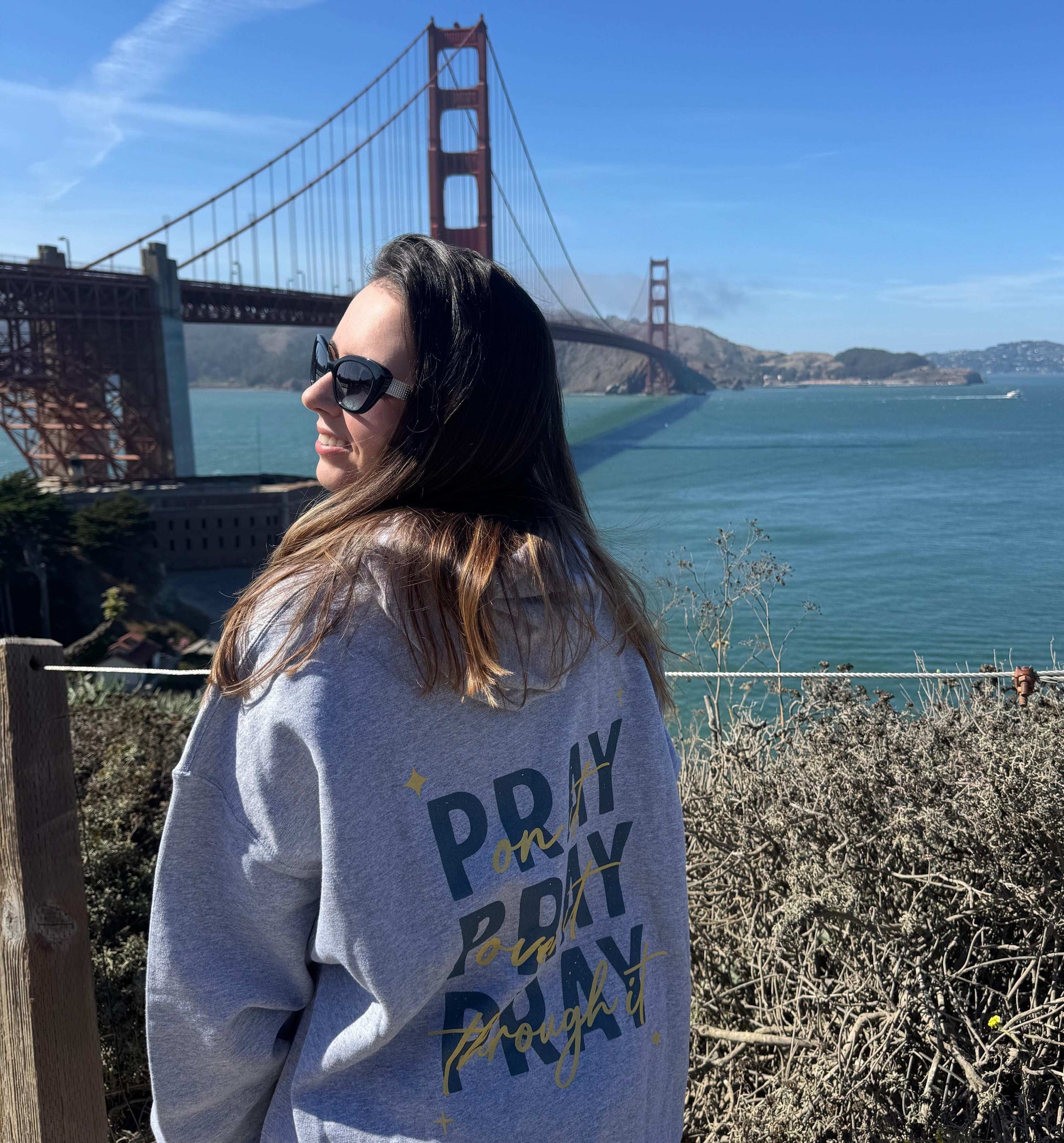 Woman wearing gray Pray On It Faith Hoodie with Golden Gate Bridge in background, promoting prayer and faith.