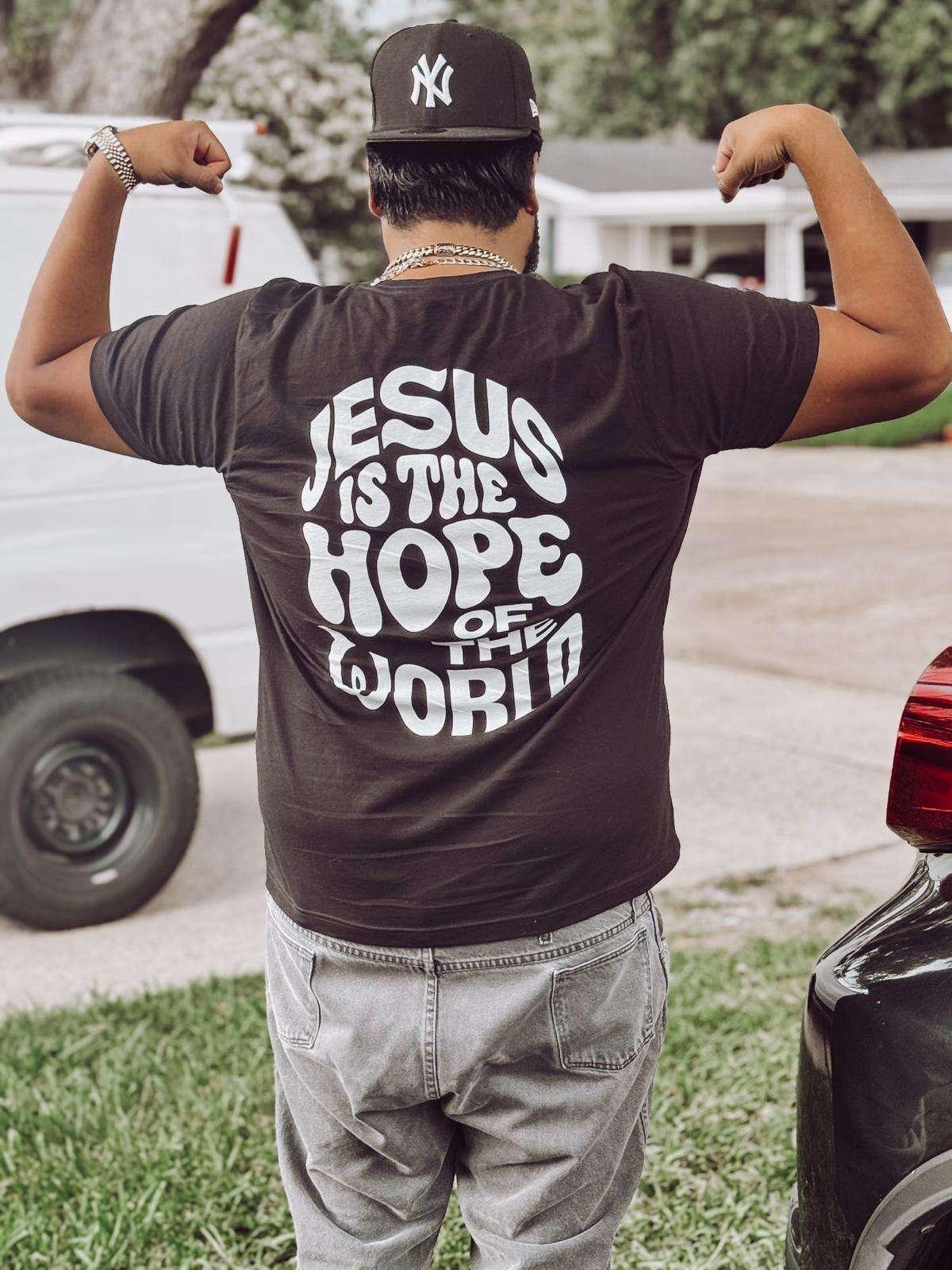 Person wearing a black t-shirt with 'Jesus is the Hope of the World' text, flexing muscles outdoors.