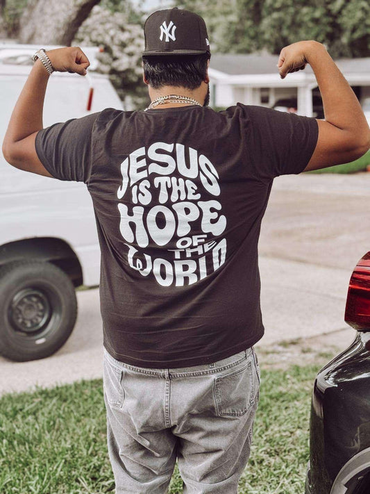 Person wearing a black t-shirt with 'Jesus is the Hope of the World' text, flexing muscles outdoors.