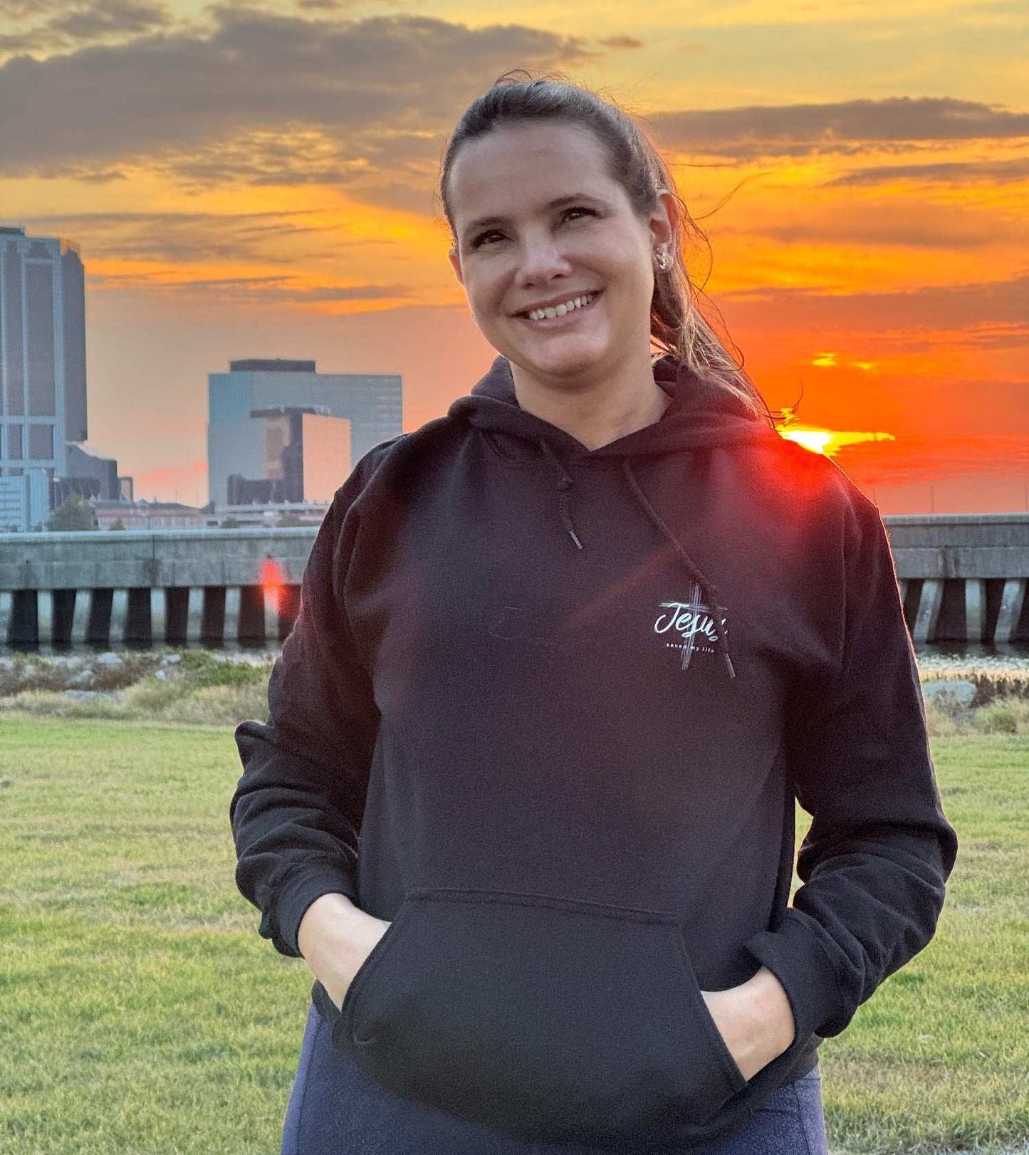 Person wearing a black hoodie with a logo, standing in front of a city skyline during sunset.