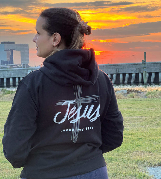 Person wearing a black hoodie with 'Jesus saved my life' text at sunset.