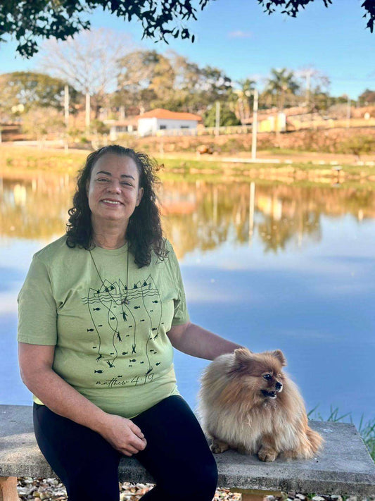 Woman wearing a green Fishers of Men Matthew 4:19 tee sitting by a lake with a small fluffy dog on a bench