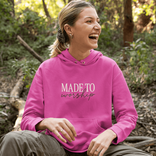 Person wearing a pink hoodie with 'MADE TO worship' text, sitting outdoors.